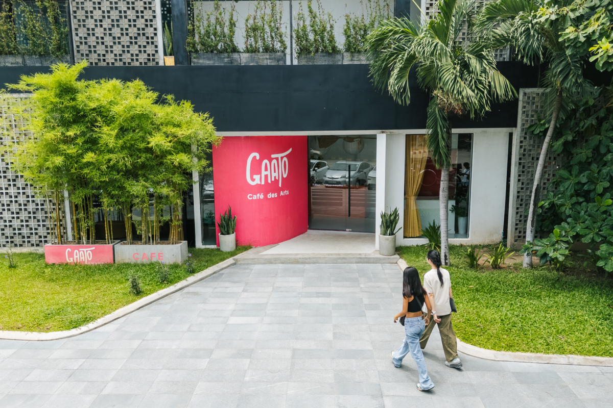 Two cafe-goers walking toward the entrance of Gaato Café des Arts, featuring a bright pink accent wall and tropical greenery.