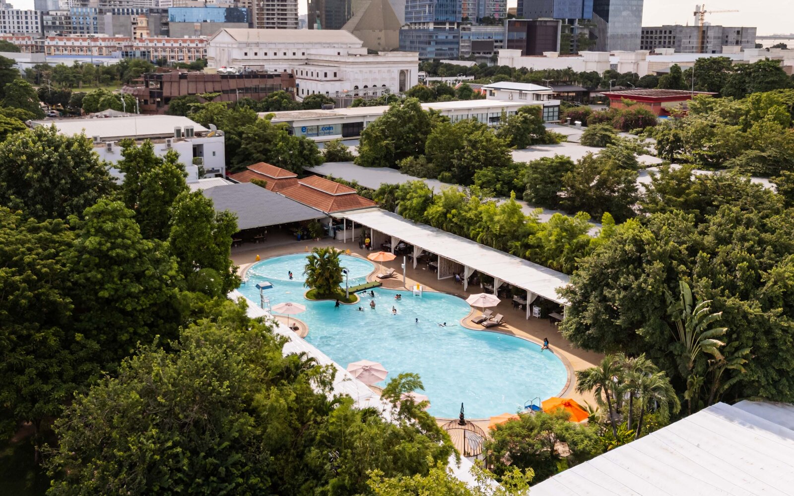 Aerial view of Dib Club swimming pool complex surrounded by trees, with city buildings in the background.