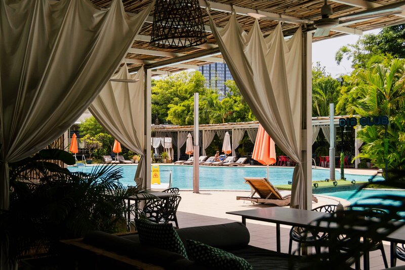 View of the outdoor pool and sun loungers framed by cabana curtains at DIB Club, Phnom Penh