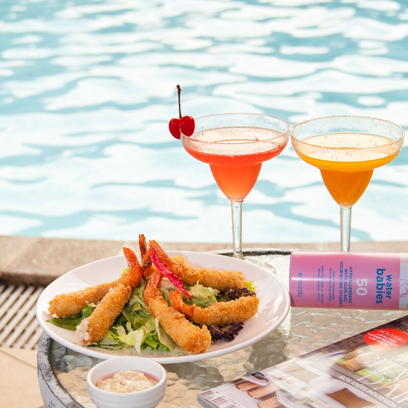 Plate of fried shrimp, two cocktails, and sunscreen spray on a poolside table, DIB Club, Phnom Penh.