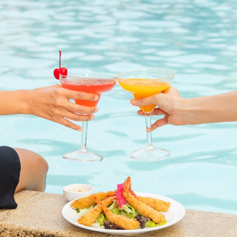 Two hands clinking cocktail glasses over fried shrimp by the swimming pool at DIB Club, Phnom Penh.