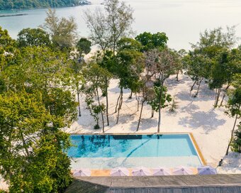 High-angle drone perspective showing the swimming pool surrounded with lush tropical trees, white sandy grounds, and the ocean at Island Center Point hotel, Koh Rong Sanloem.