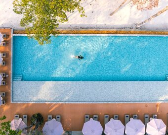 Bird's-eye view of a solo swimmer doing lap in the blue pool, framed by rows of umbrellas, wooden decking, and white sand at Island Center Point hotel, Koh Rong Sanloem.