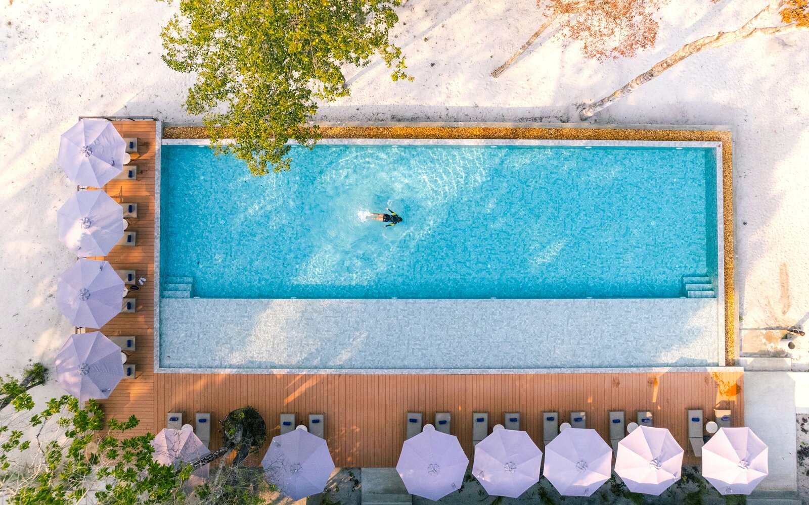 Bird's-eye view of a solo swimmer doing lap in the blue pool, framed by rows of umbrellas, wooden decking, and white sand at Island Center Point hotel, Koh Rong Sanloem.