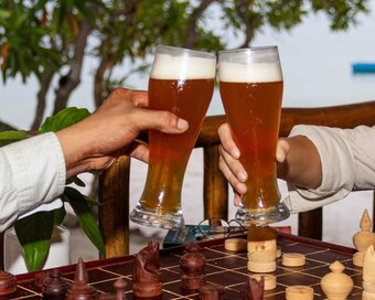 Two guests clinking glasses of craft beer over a wooden Khmer chess board with sea views in the background at Island Center Point hote, Koh Rong Sanloem.