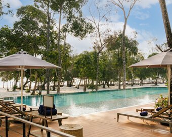 Wooden pool deck with sun loungers and umbrellas, leafy trees leading out toward the sandy shores of Island Center Point, Koh Rong Sanloem.