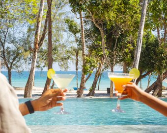 Two island-goers clinking cocktail glasses in a happy hour toast over the swimming pool and beach backdrop at Island Center Point, Koh Rong Sanloem.