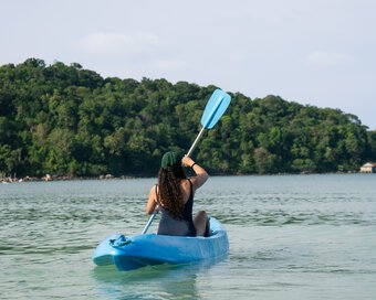 An islander paddling a kayak across ocean waters with lush green hills in the background at Island Center Point hotel, Koh Rong Sanloem.