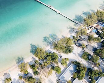Overhead drone view of turquoise water, a long wooden pier, and white sand beach lined with trees.