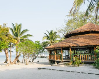 Daytime view of the round wooden beach bar nestled among palm trees on a clean white sand beach at Island Center Point, Koh Rong Sanloem.