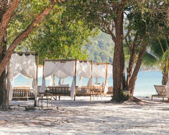 Row of shaded beach cabanas with white curtains set under trees along the sand of Saracen Bay, Koh Rong Sanloem