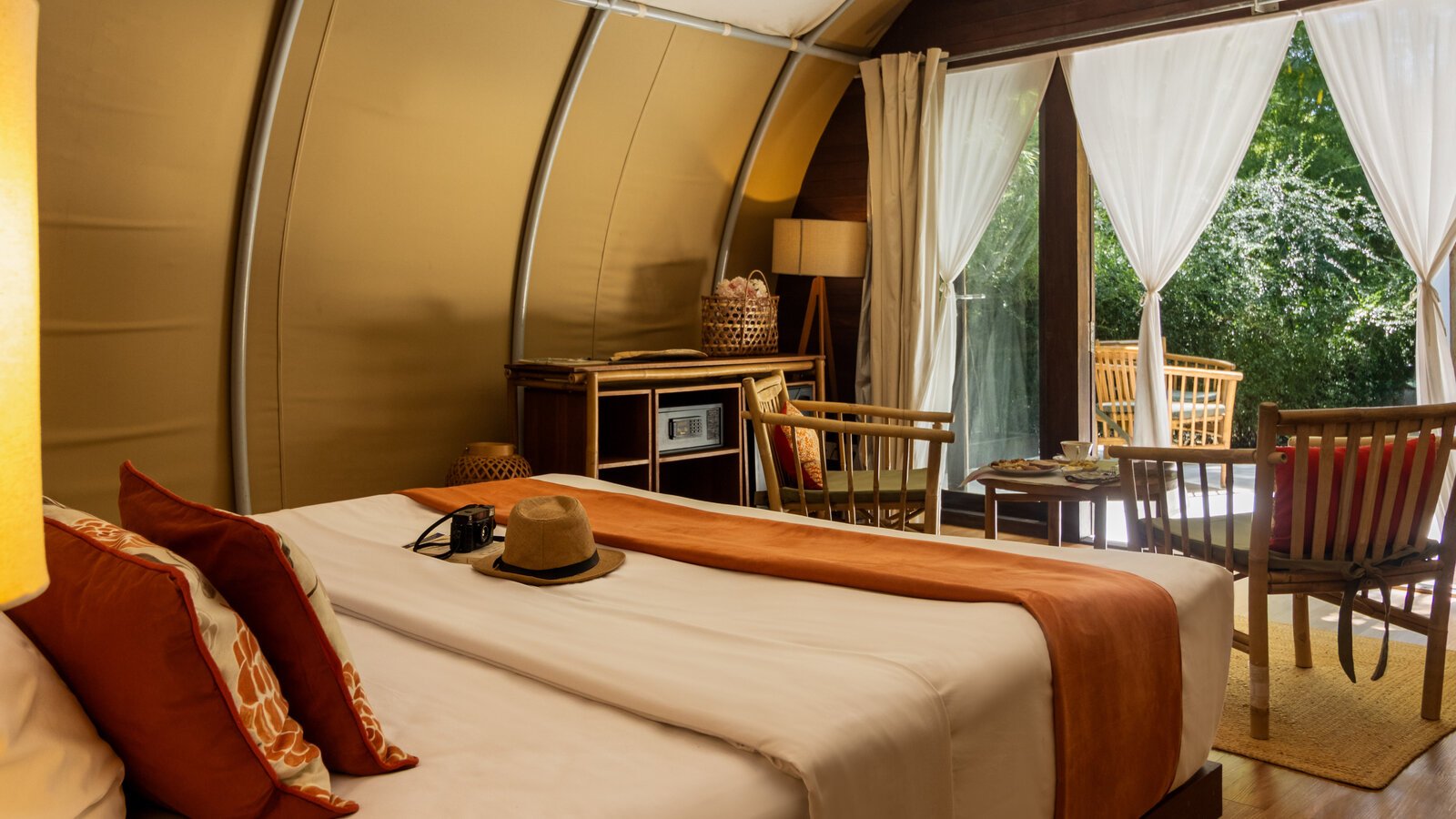 Inside a tented suite showing the bed area with orange cushions and a straw hat, with a view through open white curtains out to a garden patio with bamboo chairs.