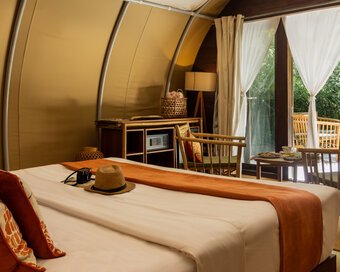 Inside a tented suite showing the bed area with orange cushions and a straw hat, with a view through open white curtains out to a garden patio with bamboo chairs.