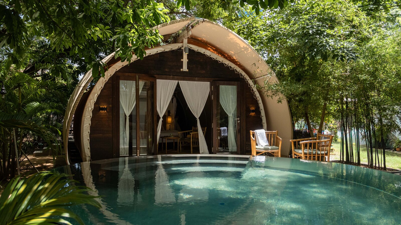 Daytime exterior view of a rounded tented wooden cabin structure (Jungloo), situated directly in front of the swimming pool surrounded by lush green trees.
