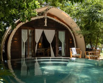 Daytime exterior view of a rounded tented wooden cabin structure (Jungloo), situated directly in front of the swimming pool surrounded by lush green trees.