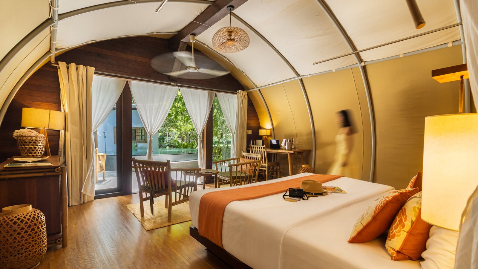 View inside the tented room featuring a bed with orange accents, wooden flooring, a ceiling fan, and large glass doors opening directly out to the pool.