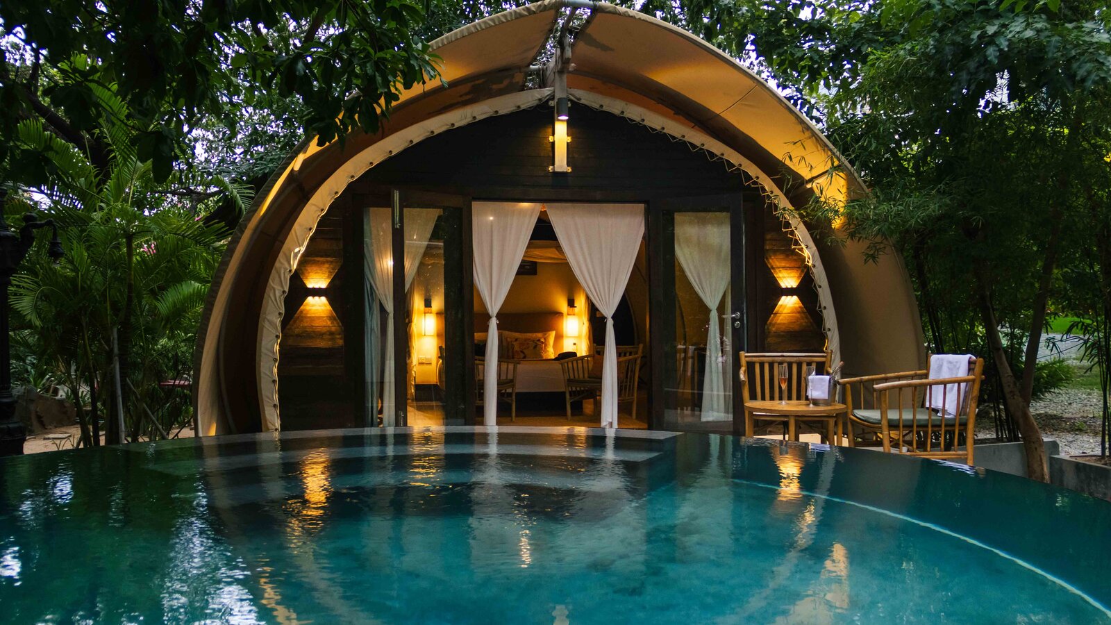 Exterior view at dusk of the rounded tented cabin showing warm indoor lighting, reflected on the surface of the plunge pool.
