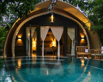 Exterior view at dusk of the rounded tented cabin showing warm indoor lighting, reflected on the surface of the plunge pool.