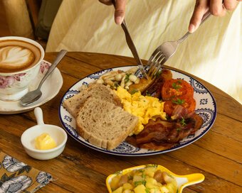 Close-up of the breakfast platter with scrambled eggs, bacon, sausage, and toast next to a cup of latte art on a wooden table.