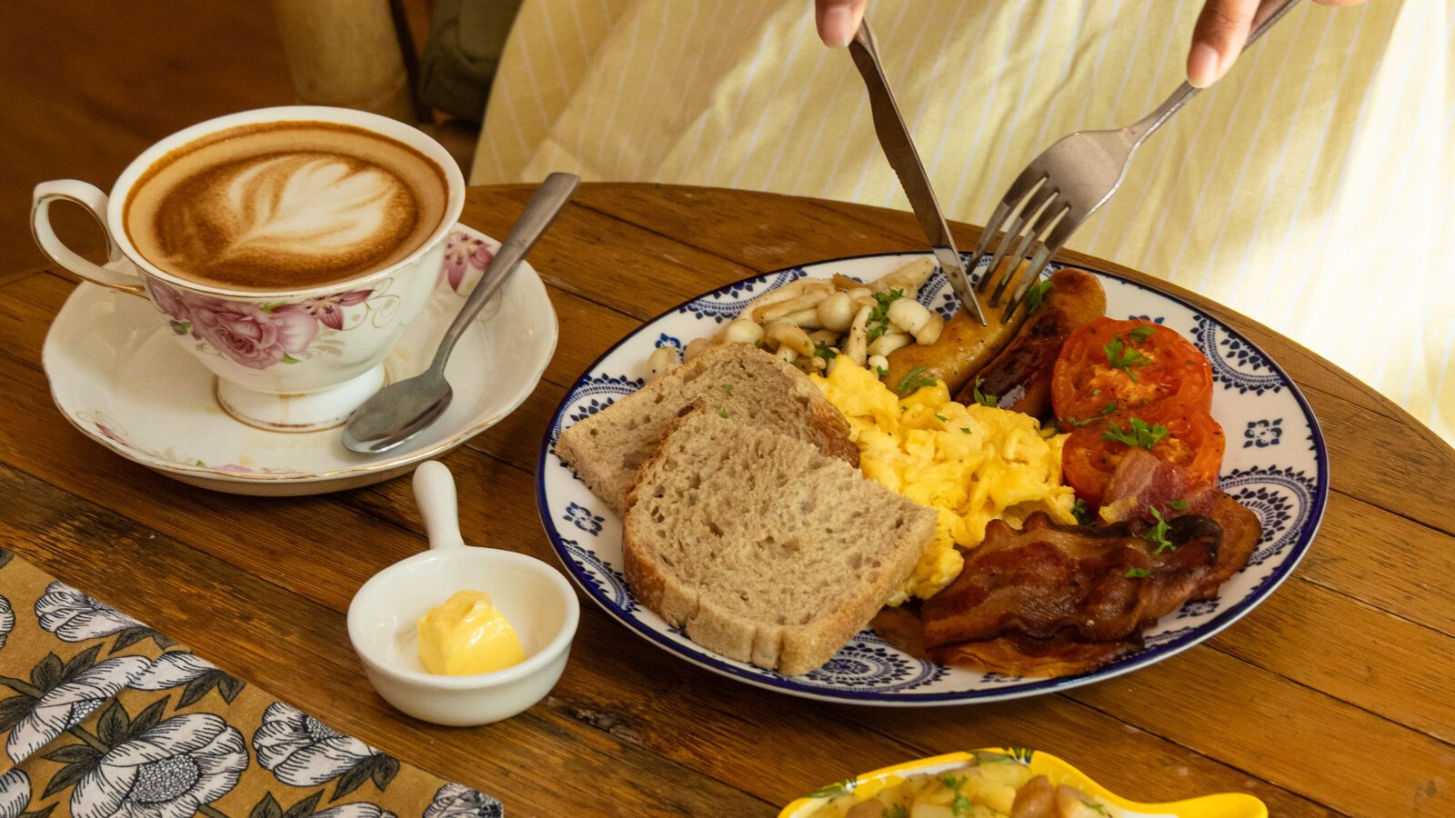 Close-up of the breakfast platter with scrambled eggs, bacon, sausage, and toast next to a cup of latte art on a wooden table.