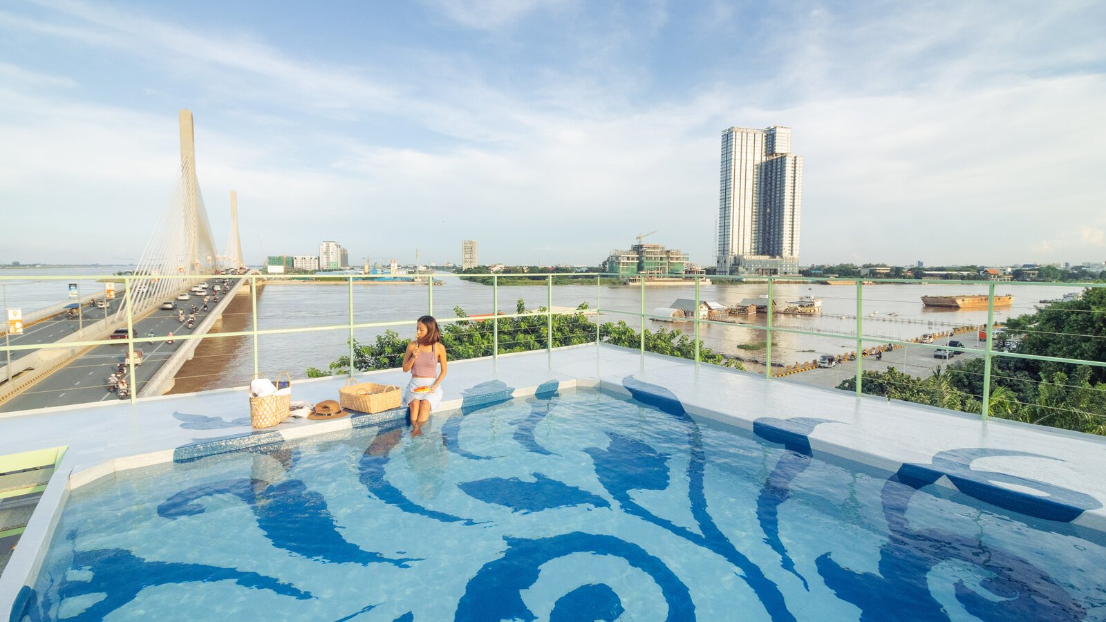 Rooftop swimming pool with a blue artwork patterned floor overlooking the bridge and river during the day.