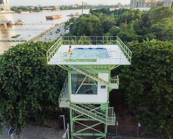 Aerial view showing the full structure of the light green water tower with a rooftop pool, set near the river and trees.