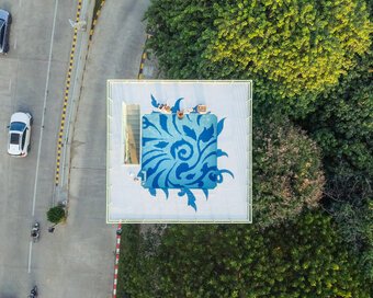 Direct top-down view of the water tower rooftop pool with blue Kbach artwork, with a road on the left and dense trees on the right.