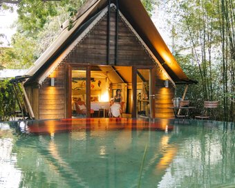Evening view of Park Jungloo unit (Jungle Igloo) made of wood, illuminated by warm lights, with a woman seated inside the glass doors, looking out over the private plunge pool.