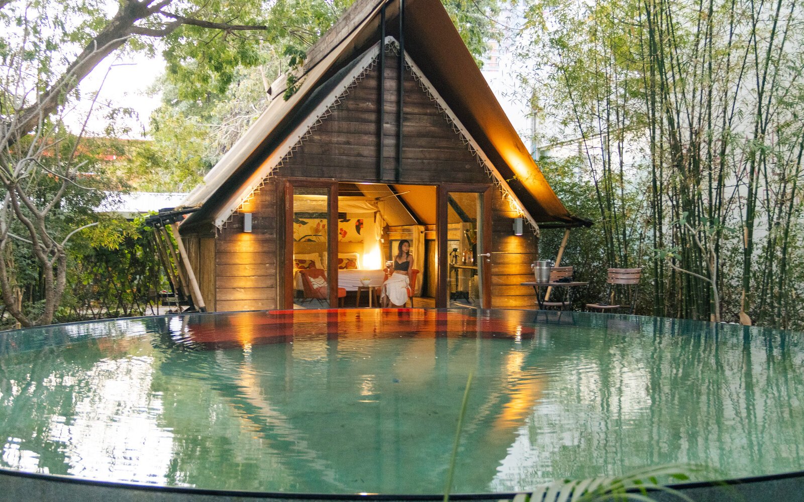 Evening view of Park Jungloo unit (Jungle Igloo) made of wood, illuminated by warm lights, with a woman seated inside the glass doors, looking out over the private plunge pool.