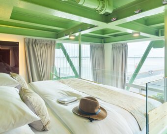 Interior view of the bed featuring traveller items, framed by industrial green steel and large glass windows overlooking the river and road.