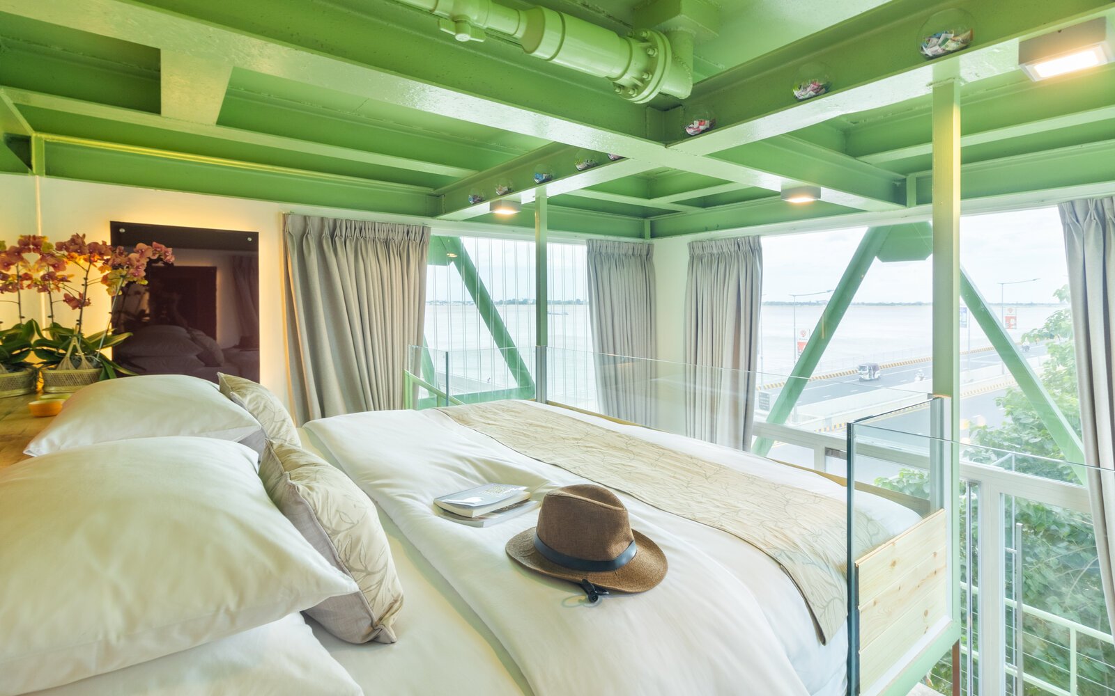 Interior view of the bed featuring traveller items, framed by industrial green steel and large glass windows overlooking the river and road.
