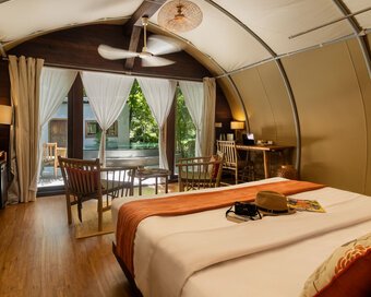 Interior view of Jungloo (Jungle Igloo) unit, showing a bed with an orange runner, wooden seating, and large glass doors opening to the outside.