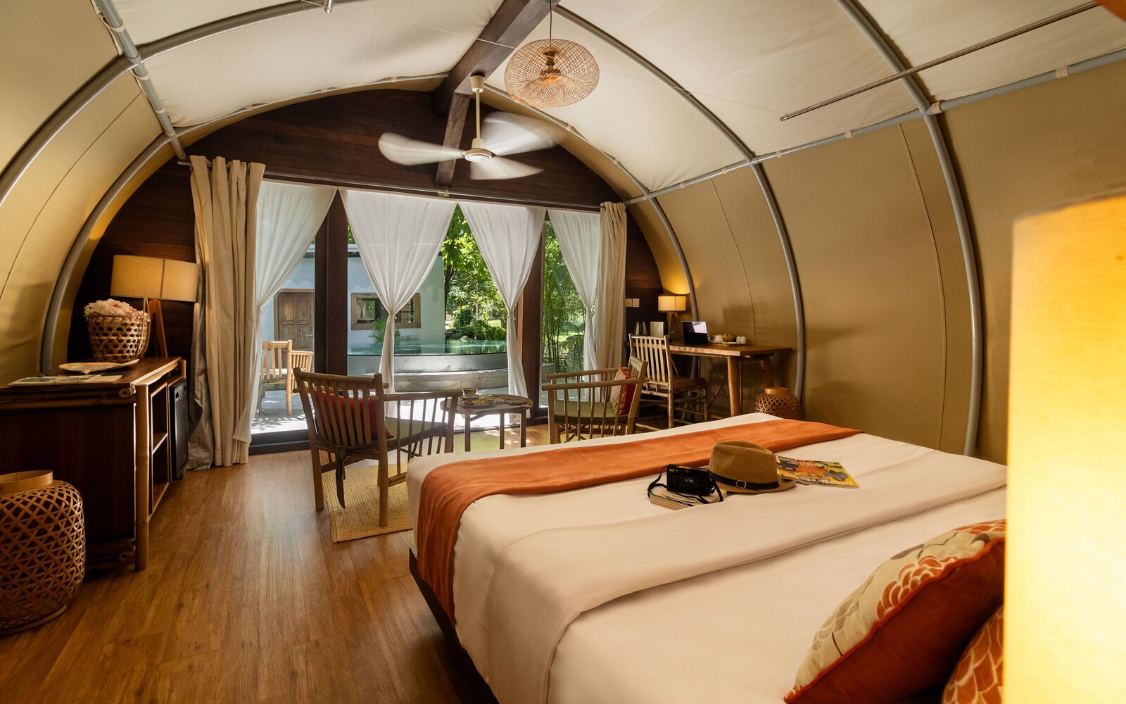 Interior view of Jungloo (Jungle Igloo) unit, showing a bed with an orange runner, wooden seating, and large glass doors opening to the outside.