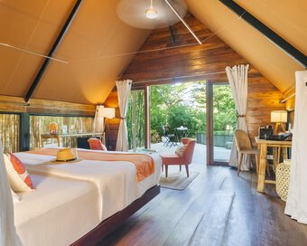 Inside Jungloo unit showing the bed with an orange runner, polished wood floors, and large glass doors leading to the green outdoor deck.