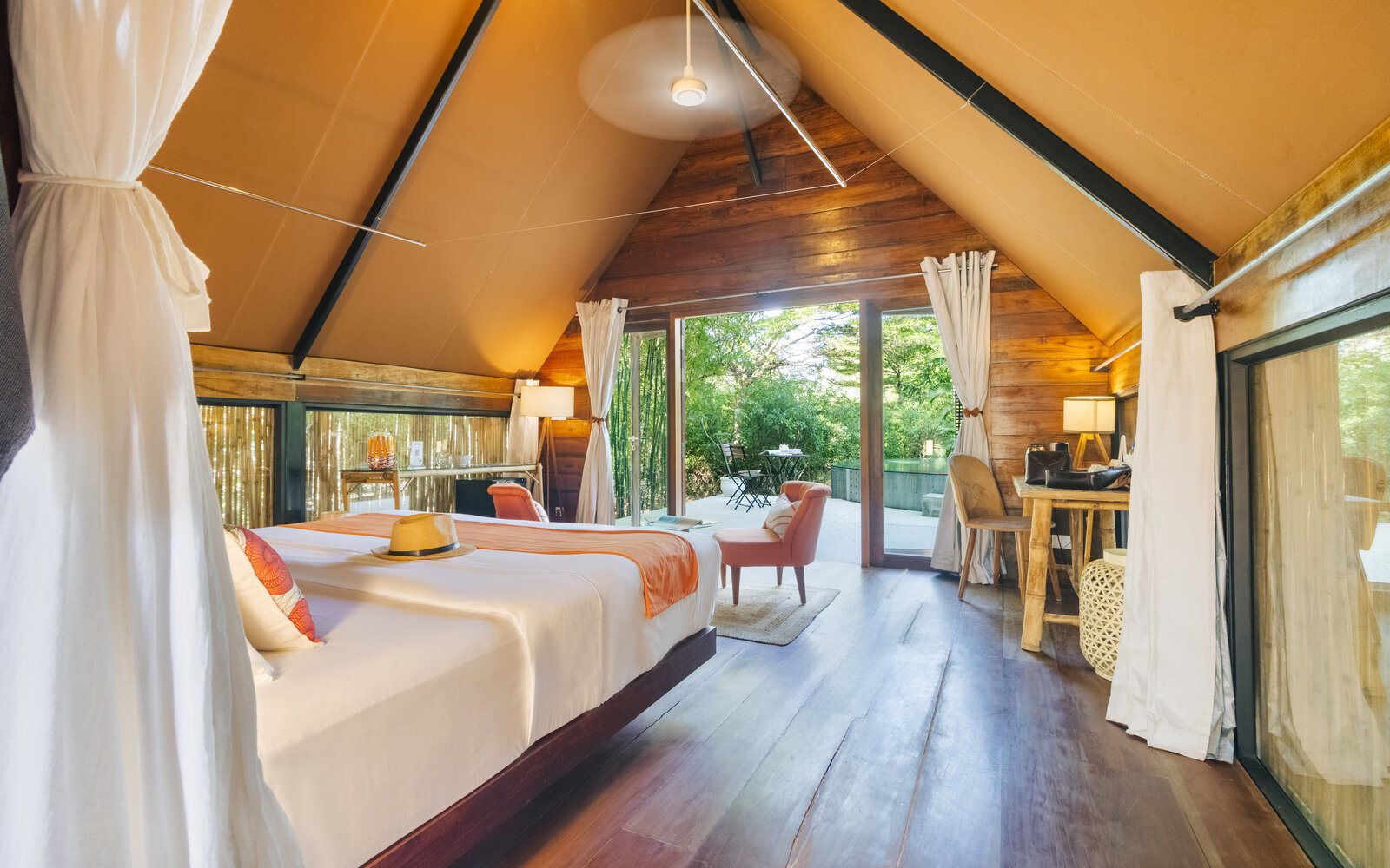 Inside Jungloo unit showing the bed with an orange runner, polished wood floors, and large glass doors leading to the green outdoor deck.