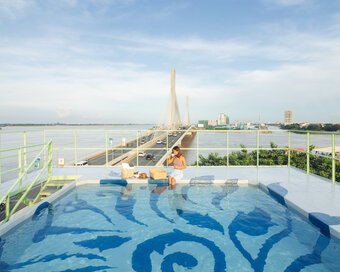 Daylight view of the rooftop pool featuring blue Kbach Cambodian artwork, with guests sitting on the edge, with a background of the bridge over a wide river.