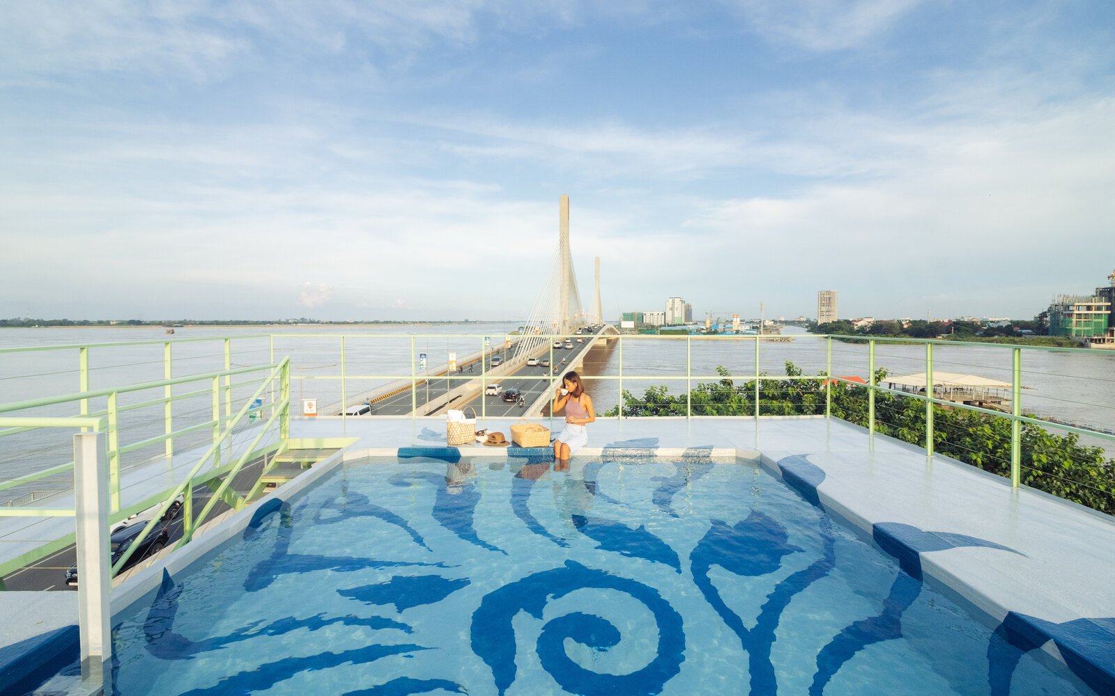 Daylight view of the rooftop pool featuring blue Kbach Cambodian artwork, with guests sitting on the edge, with a background of the bridge over a wide river.