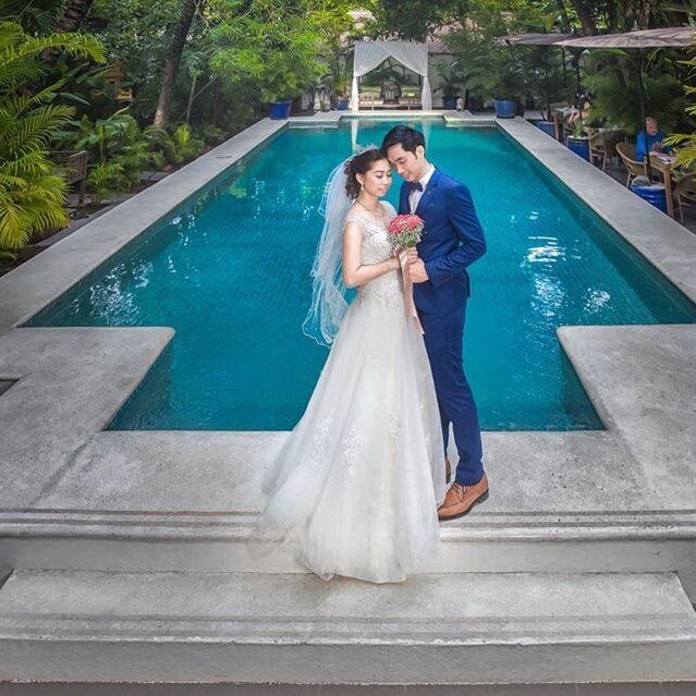 Bride and groom pause for a photo on the pool steps with palm trees and a covered cabana behind them.