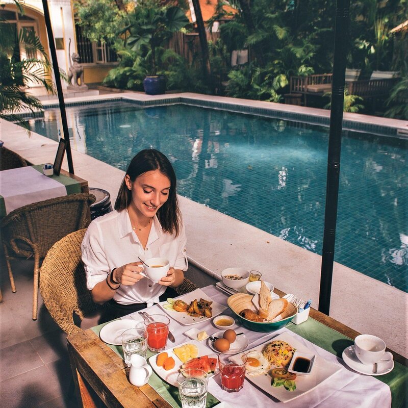 Breakfast is served poolside as a guest lifts her cup, with the lush pool behind.