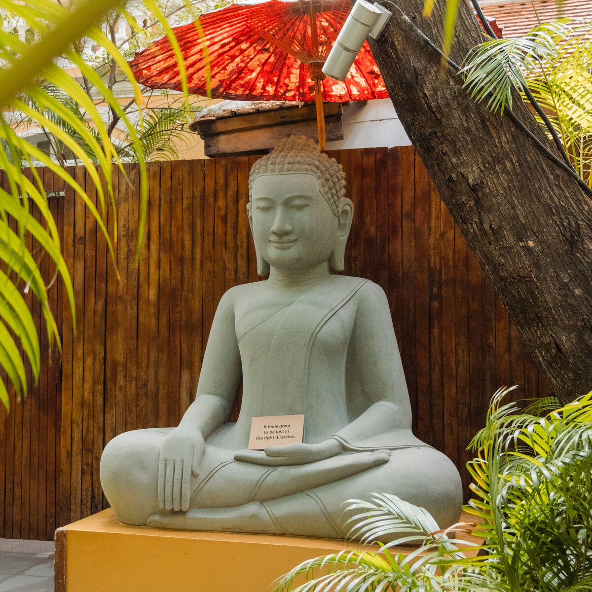 Stone Buddha statue sits in the garden under a red umbrella, a small note tucked in its hand.