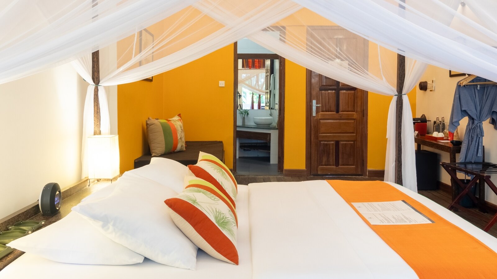 Queen bed with mosquito net, seating area, guest bathrobe, and bathroom entrance in a Double Deluxe Room at Pavilion Phnom Penh