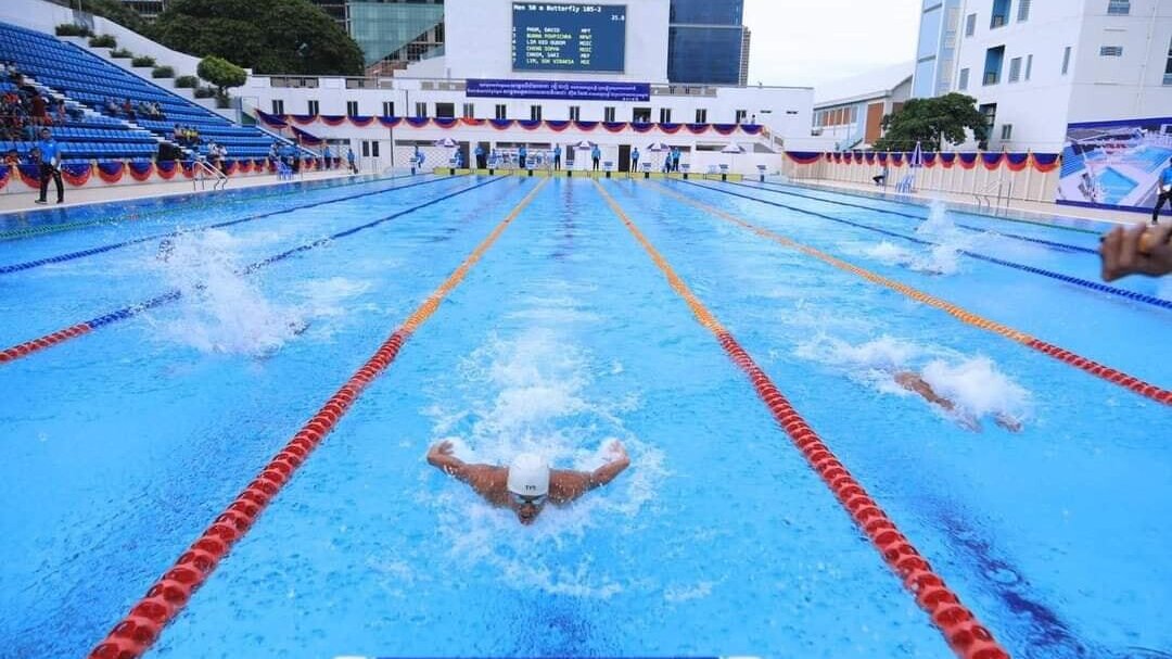 Competitive swimming lanes at the National Olympic Stadium pool during a race event, framed by spectator seating and scoreboards.