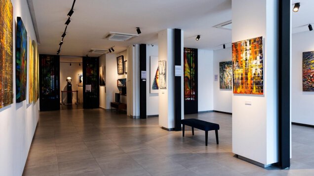 Bright gallery space displaying contemporary paintings across multiple exhibition rooms, with open walkways connecting the collection.