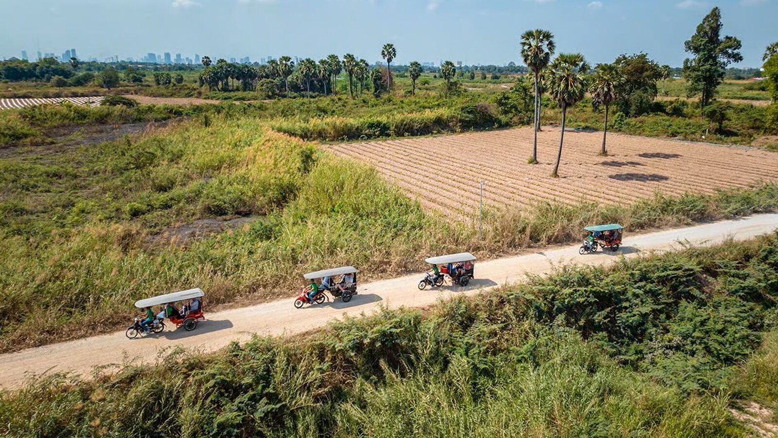 Line of tuk tuks follows a rural road through open field landscapes.