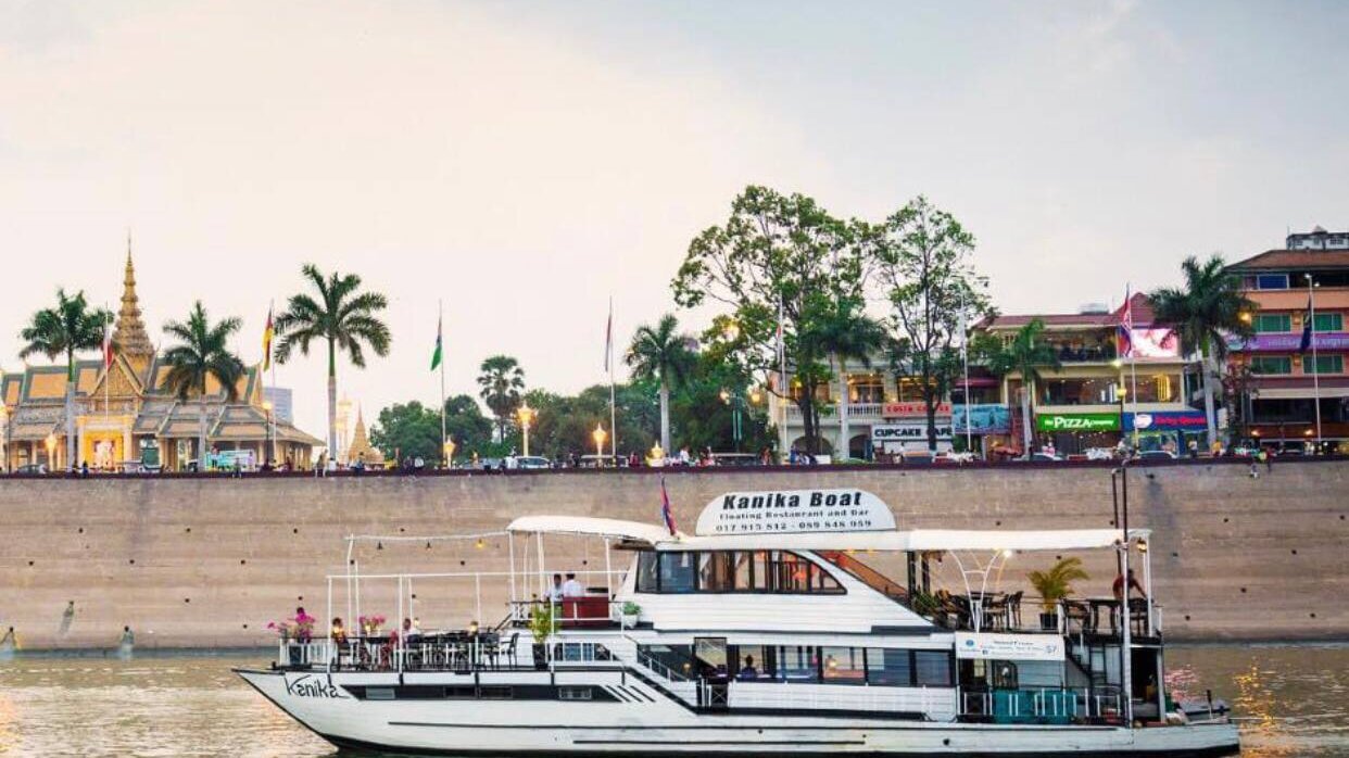 Guests gather on an open-air river cruise deck at sunset with views toward Phnom Penh's riverfront and the Royal Palace area.