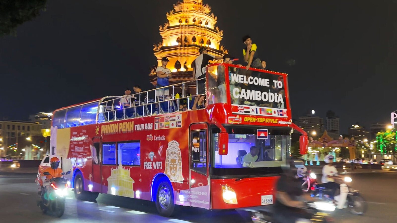 An open-top sightseeing bus passes illuminated Independence Monument at evening tour through Phnom Penh.