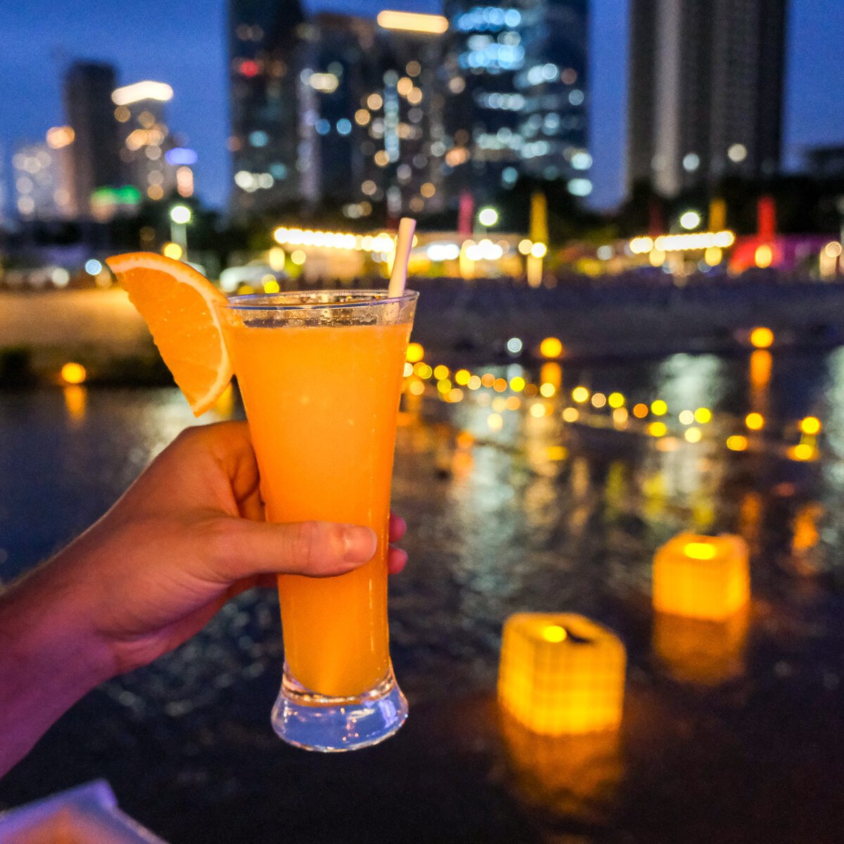 Citrus-garnished evening cocktail held above illuminated riverside waters with city lights beyond.