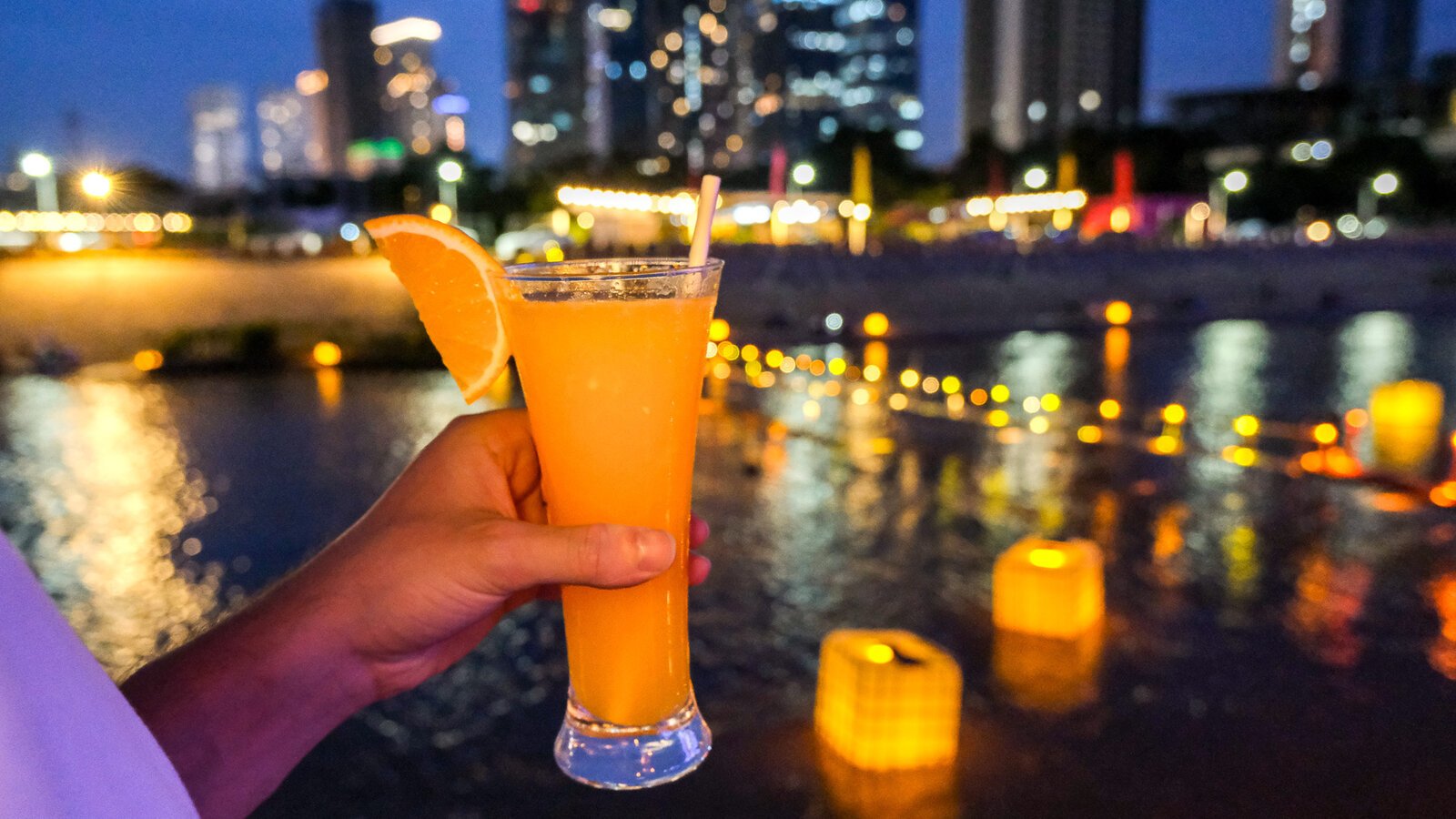 Citrus-garnished evening cocktail held above illuminated riverside waters with city lights beyond.