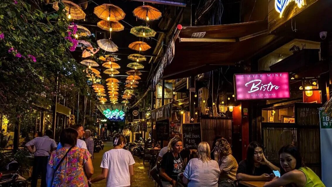 Visitors gather beneath hanging umbrellas and illuminated signs along a lively alleyway at night.