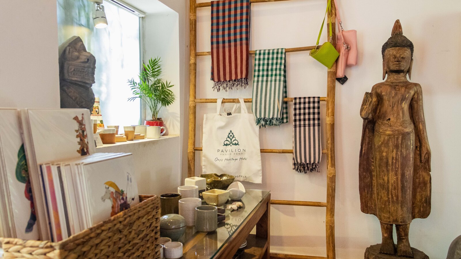 Display of Cambodian textiles, ceramic pieces, tote bags, and decorative items beside a Cambodian carved Buddha.
